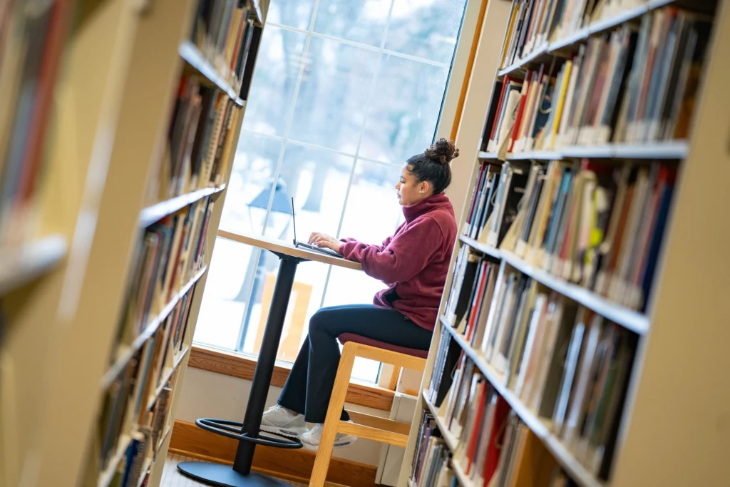 A person sits at a high table between bookshelves in a library, working on student publications on a laptop while facing a large window with a snowy scene outside.