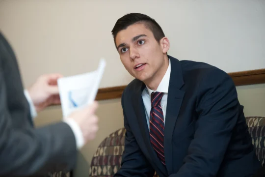 A young man in a dark suit and red striped tie sits on a patterned chair, attentively listening to someone off-camera discuss funding for real-world learning while holding a sheet of paper.