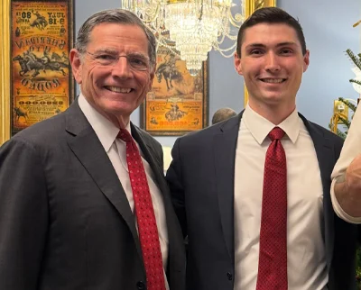 Two men wearing dark suits, white shirts, and red ties stand smiling together indoors. Behind them are antique posters, a chandelier, and part of a decorated Christmas tree.