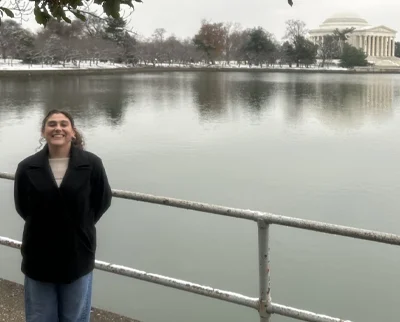A person smiles while standing by a metal railing in front of a calm body of water, with the Jefferson Memorial and snow-dusted trees visible in the background.