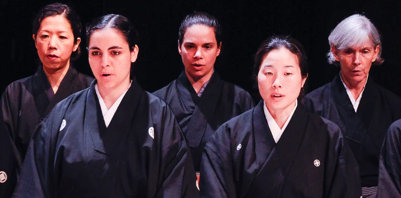 Five women dressed in traditional black kimono stand in two rows, facing forward against a dark background, with serious and focused expressions.