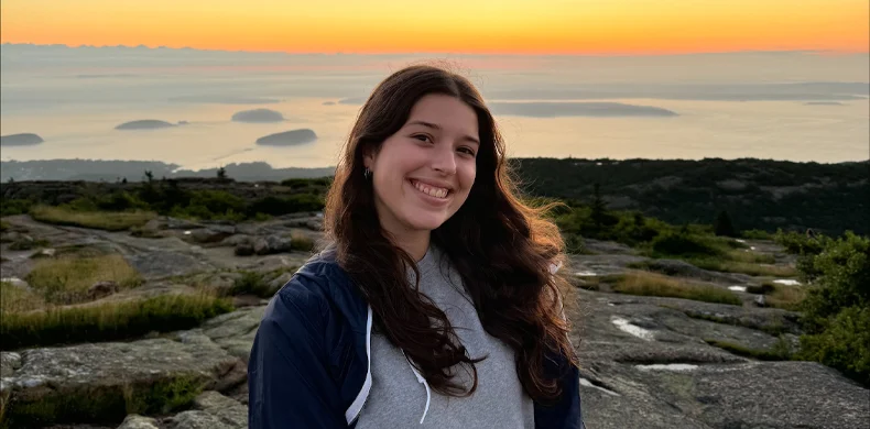 A young woman with long brown hair smiles at the camera while standing on rocky terrain, with a scenic sunset and islands visible in the background.