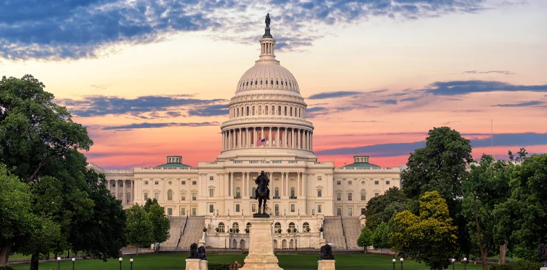 The United States Capitol building in Washington, D.C., is shown at sunset with a colorful sky. Green trees and a statue are in the foreground.
