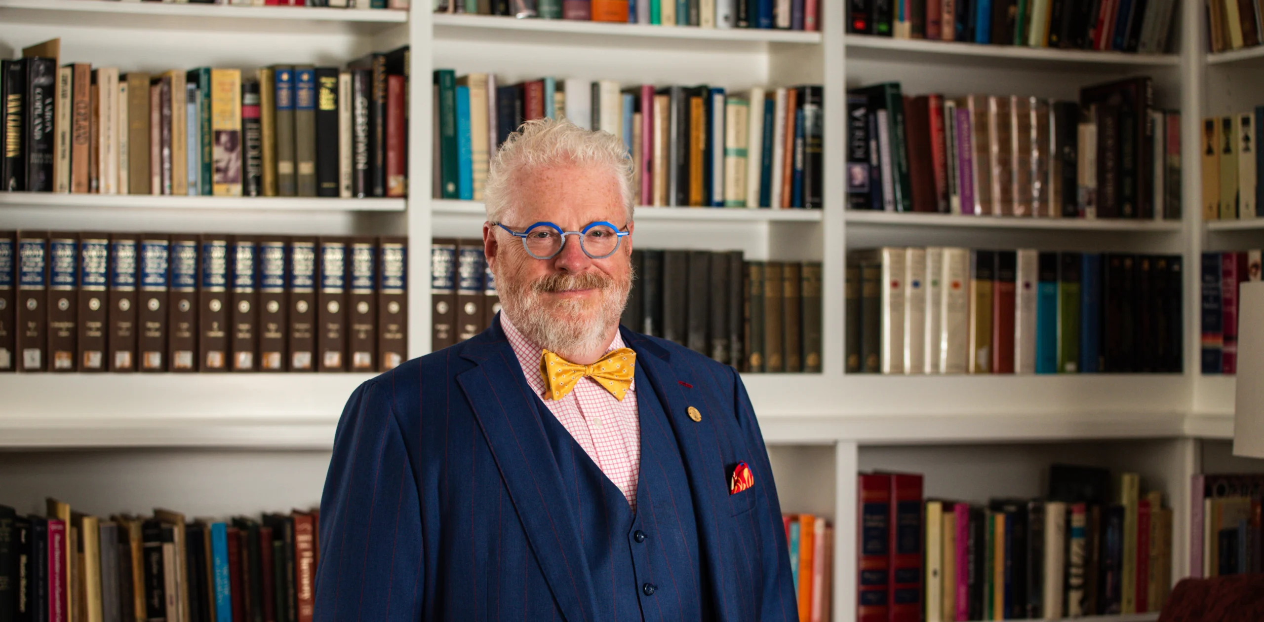 An older man with white hair and beard, wearing blue glasses, a yellow bow tie, and a blue suit, stands in front of bookshelves filled with books.